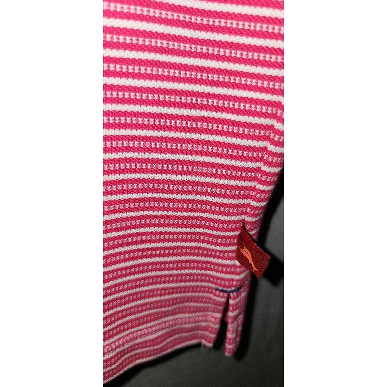 Puede incluir: Primer plano de un polo a rayas rosas y blancas. La camisa tiene un patr&oacute;n de punto texturizado y una peque&ntilde;a etiqueta roja con un logotipo. La camisa tiene un dobladillo inferior dividido.