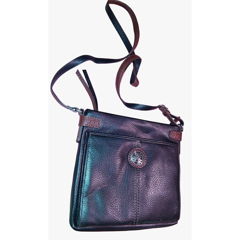 Puede incluir: Un bolso bandolera de cuero texturizado en color marr&oacute;n oscuro con una correa larga y ajustable, un bolsillo exterior frontal y un emblema circular de metal plateado centrado en la solapa delantera con detalles de volutas en relieve.