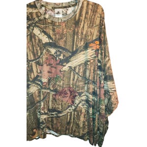 May include: Long-sleeved camouflage shirt in shades of brown, green, and tan. The shirt features a crew neck and a pattern of leaves and branches. The Mossy Oak Break-Up logo is visible on the right sleeve.