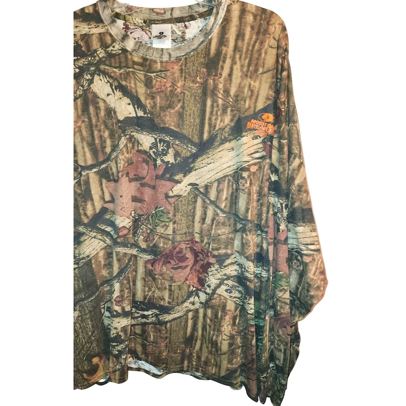 May include: Long-sleeved camouflage shirt in shades of brown, green, and tan. The shirt features a crew neck and a pattern of leaves and branches. The Mossy Oak Break-Up logo is visible on the right sleeve.