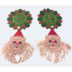May include: A pair of decorative Santa Claus ornaments featuring felt faces with sequined hats and beards made of light-colored fringe. Each Santa is topped with a green felt wreath adorned with red sequins and green holly leaf accents.