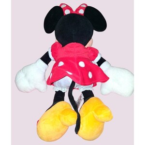 May include: A plush Minnie Mouse doll, viewed from the back, features a red dress with white polka dots, a matching bow, and large yellow shoes. The doll has black ears, a black tail, and white gloves.