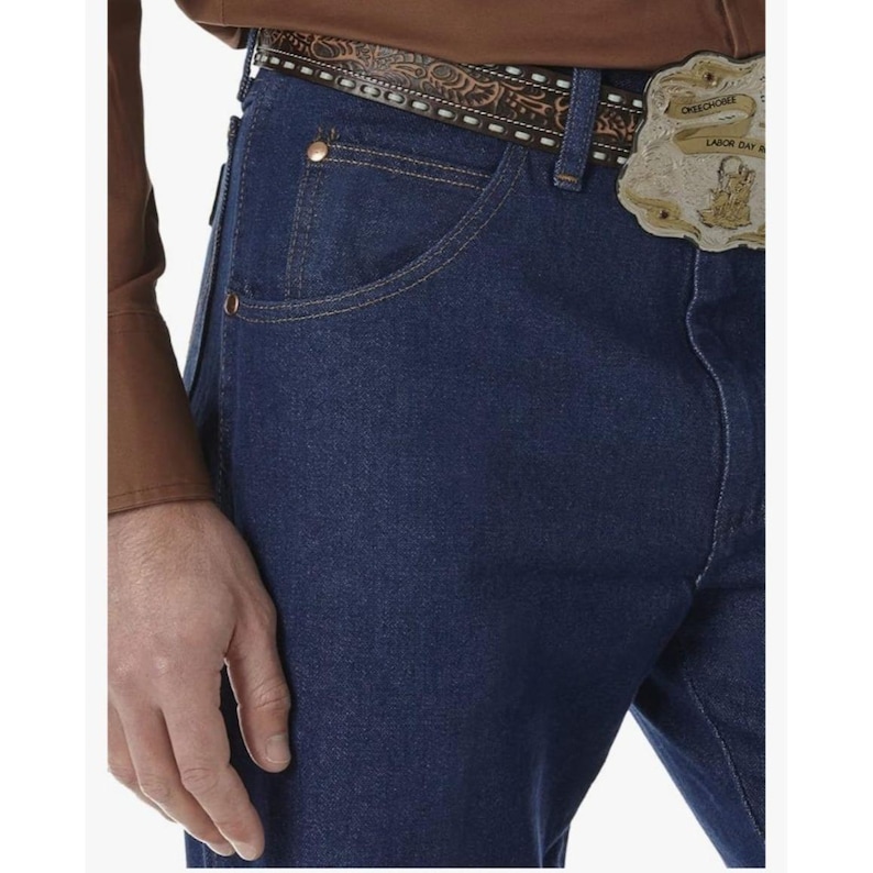 May include: Close-up of dark blue denim jeans with a brown leather belt featuring a decorative pattern and a gold-colored belt buckle. The jeans have classic five-pocket styling and copper-colored rivets.