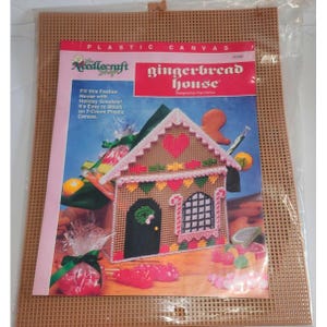 May include: A Needlecraft plastic canvas kit for a gingerbread house. The kit includes a 7-count plastic canvas, instructions, and a pattern for a gingerbread house with candy cane trim and a heart-shaped window. The image shows a completed gingerbread house filled with candy.