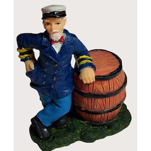 May include: A nautical-themed figurine of a sailor leaning against a wooden barrel. The sailor wears a blue jacket with gold stripes, blue pants, a white hat, and a red bow tie. The barrel is brown with visible wood grain, and the base is green.