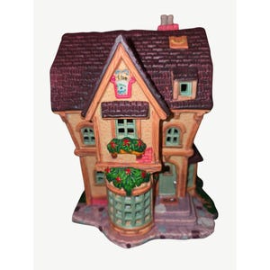 May include: A ceramic miniature house figurine featuring a dark purple shingled roof, tan walls, and a green lattice bay window. The structure displays a sign reading The Flower Pot, green window boxes with red flowers, and a small chimney on top.