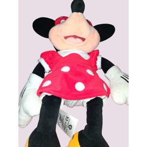 May include: A plush Minnie Mouse doll. The doll has a red dress with white polka dots, white gloves, and black legs. Minnie's face is pink with a large smile, and she wears a red bow with white polka dots. The doll has black ears and yellow shoes.