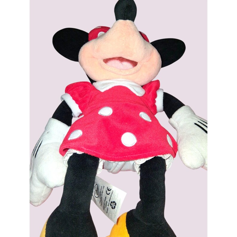 May include: A plush Minnie Mouse doll. The doll has a red dress with white polka dots, white gloves, and black legs. Minnie's face is pink with a large smile, and she wears a red bow with white polka dots. The doll has black ears and yellow shoes.