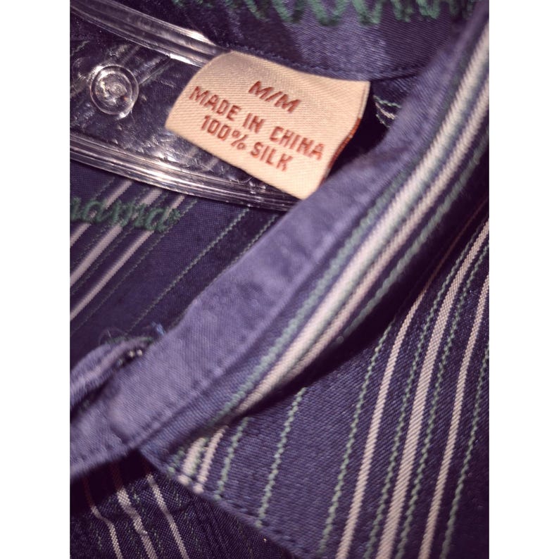 May include: A dark blue button-down shirt with thin vertical stripes in white and green. The collar features a beige tag with the text M/M, MADE IN CHINA, and 100% SILK. The shirt is displayed on a clear plastic hanger, showing the fabric texture.