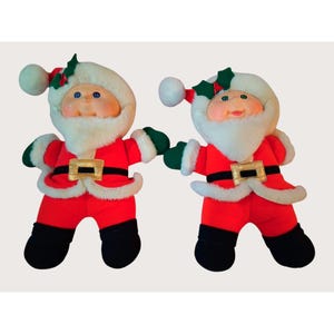Può includere: Due bambole di peluche di Babbo Natale. Ogni bambola indossa un abito rosso con cintura nera e fibbia dorata, stivali neri e guanti verdi. Hanno barbe bianche, guance rosee e occhi blu. Ogni bambola ha un cappello bianco con finiture rosse e agrifoglio.