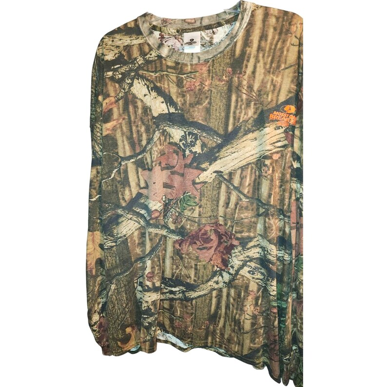 May include: Long-sleeved camouflage shirt with a crew neck. The shirt features a brown, green, and tan pattern with floral accents. The shirt has a tag on the neckline with the brand name. The fabric appears to be lightweight.