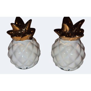 Puede incluir: Un par de saleros y pimenteros de cerámica con forma de piña. Cada pieza presenta un cuerpo blanco texturizado con patrón de diamantes y una parte superior lisa y reflectante de tono dorado que representa las hojas.