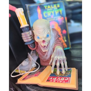 May include: A Crypt Keeper figure holding a yellow candle, featuring a pale, wrinkled face with blue eyes and white hair. The figure wears a brown robe and reaches over an open book with the text Tales from the Crypt printed on the cover.