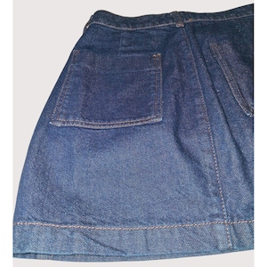 Pode incluir: Close-up de uma saia jeans em lavagem m&eacute;dia. A saia apresenta um grande bolso quadrado na lateral e costuras vis&iacute;veis em uma cor contrastante. A bainha tamb&eacute;m &eacute; costurada. A saia &eacute; um estilo cl&aacute;ssico.