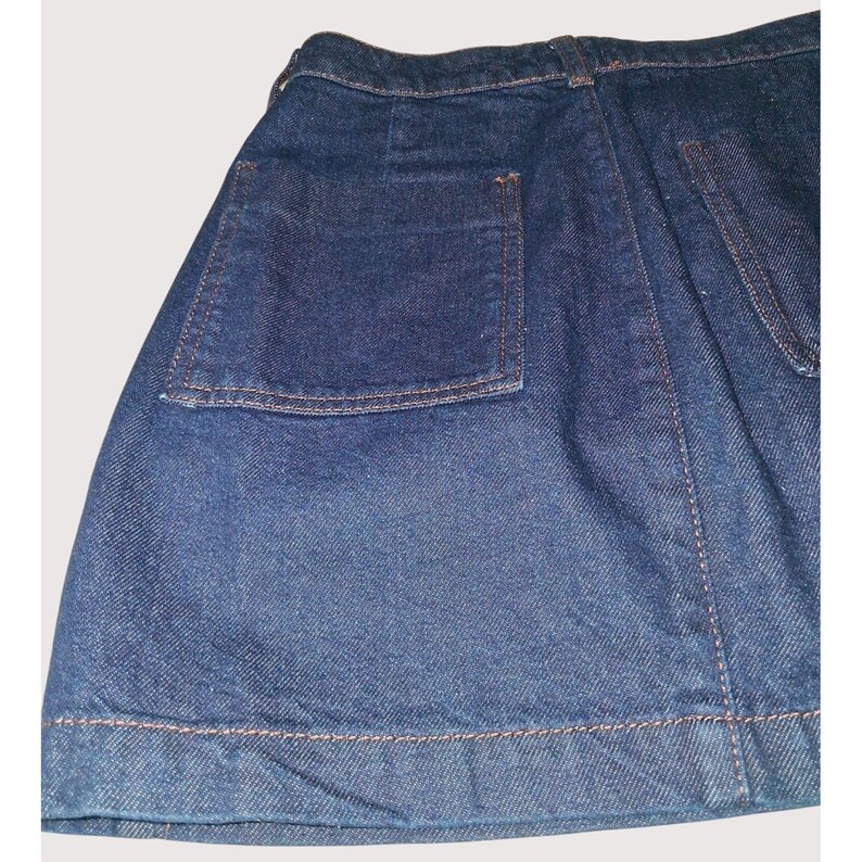 Pode incluir: Close-up de uma saia jeans em lavagem m&eacute;dia. A saia apresenta um grande bolso quadrado na lateral e costuras vis&iacute;veis em uma cor contrastante. A bainha tamb&eacute;m &eacute; costurada. A saia &eacute; um estilo cl&aacute;ssico.