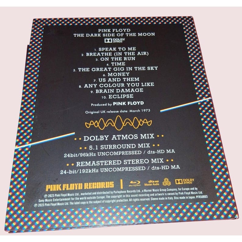 May include: The back cover of the Pink Floyd The Dark Side of the Moon album features a black background with a white tracklist, Dolby Atmos logo, and technical audio specifications. A yellow waveform graphic sits above the mix details.