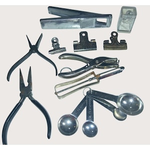 May include: Assortment of vintage metal tools and kitchen utensils. Includes pliers, paper clips, a garlic press, a hole punch, a whisk, measuring spoons, and a small glass container. The tools are arranged on a light background.