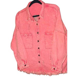 May include: A coral-colored denim jacket with a button-up front, two chest pockets, and distressed detailing. The jacket has long sleeves with buttoned cuffs and a frayed hemline. It is hanging on a black hanger.