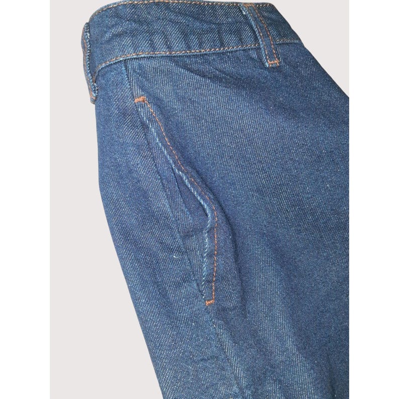 Pode incluir: Close-up de uma se&ccedil;&atilde;o de cal&ccedil;as de ganga azul escuras. A imagem destaca o bolso e a cintura, com costuras laranja vis&iacute;veis. O tecido tem uma apar&ecirc;ncia texturizada, e a est&eacute;tica geral &eacute; casual e cl&aacute;ssica.