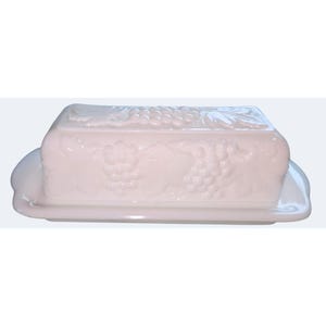 May include: A rectangular white milk glass butter dish featuring a raised, embossed pattern of grape clusters and vine leaves on the lid. The base has a smooth, slightly curved rim, and the entire piece has a glossy, opaque finish.