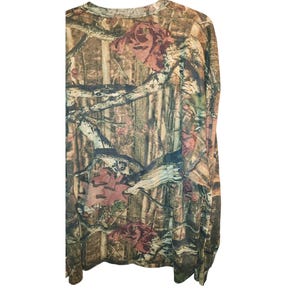May include: Long-sleeved camouflage shirt with a crew neck. The shirt features a brown, green, and tan camouflage pattern with accents of pink and red floral designs. The fabric appears to be lightweight and suitable for outdoor activities.