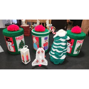 May include: Five handmade Christmas-themed decorations, including three green and white containers with red pom-poms on top, a white crocheted Christmas tree, and a white plastic bell ornament. The containers have embroidered designs of a snowman, a Christmas tree, and a nutcracker.