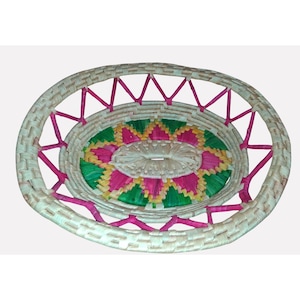 May include: Oval woven basket with a decorative floral pattern in the center. The basket features a natural beige color with pink accents and a pink zig-zag pattern around the rim. The interior design includes green, yellow, and pink colors.