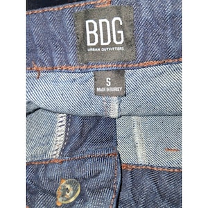 Pode incluir: Close-up de cal&ccedil;as de ganga azuis com fecho de bot&atilde;o. As cal&ccedil;as de ganga t&ecirc;m uma etiqueta preta que diz "BDG URBAN OUTFITTERS" e uma etiqueta mais pequena que diz "S MADE IN TURKEY".