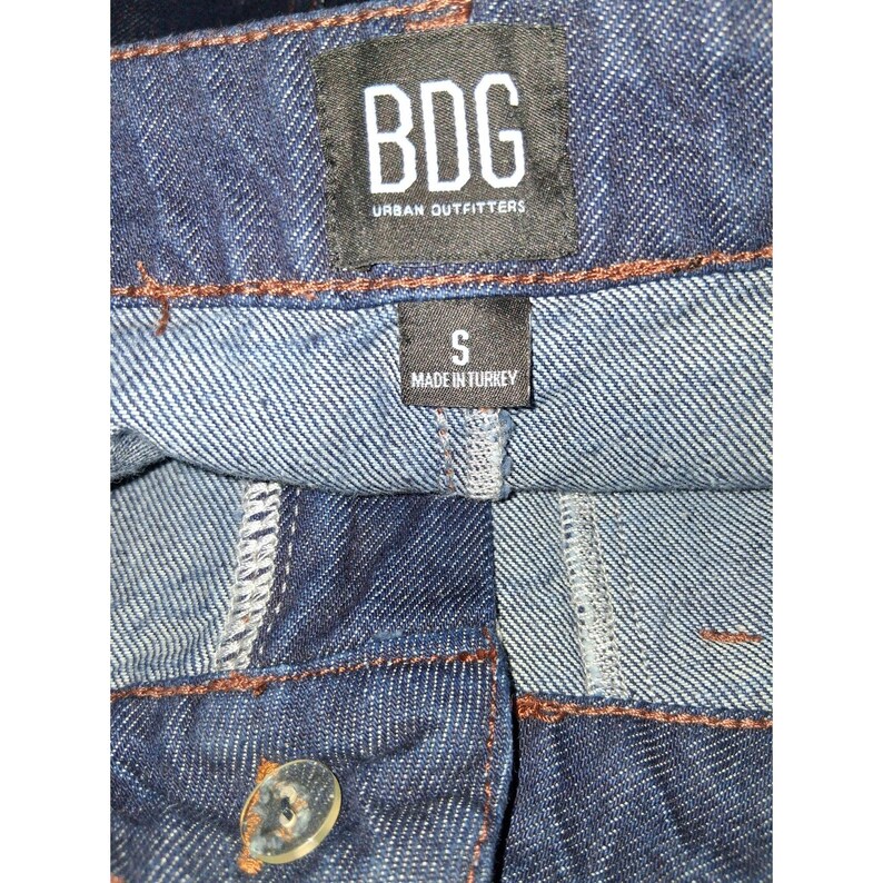 Pode incluir: Close-up de cal&ccedil;as de ganga azuis com fecho de bot&atilde;o. As cal&ccedil;as de ganga t&ecirc;m uma etiqueta preta que diz "BDG URBAN OUTFITTERS" e uma etiqueta mais pequena que diz "S MADE IN TURKEY".