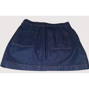 Pode incluir: Uma saia jeans de lavagem escura com dois bolsos traseiros. A saia tem um design cl&aacute;ssico com bainha reta e costuras vis&iacute;veis. O tecido parece ser um jeans de peso m&eacute;dio, adequado para uso casual.