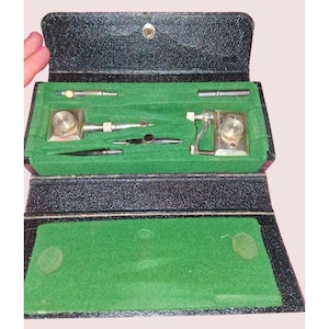 May include: A black textured case with a green velvet interior containing a set of silver-toned metal drafting instruments. The kit includes various precision tools, including compass components and specialized drawing attachments, arranged in molded slots.