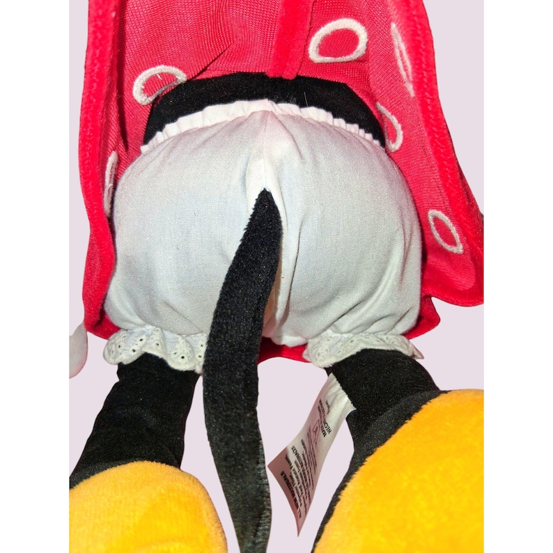 May include: A plush toy featuring a red cape with white polka dots, white bloomers with lace trim, and black legs with yellow shoes. The toy has a black tail and a tag.