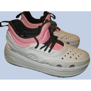 May include: Pair of white and pink athletic shoes with black laces. The shoes have a perforated design on the toe and sides, and a thick sole. The shoes have a pull tab on the back and the brand logo on the side.