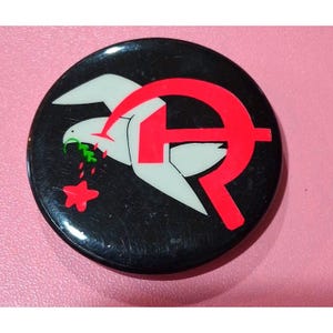 Könnte beinhalten: Ein runder, schwarzer Button mit einem weißen Vogeldesign, einem roten Hammer und Sichel sowie einem roten Stern. Der Vogel hat einen grünen Olivenzweig im Schnabel und rote Tropfen fallen aus ihm heraus.