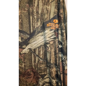 May include: Long-sleeved camouflage shirt with a pattern of brown, green, and tan. The shirt features the "Mossy Oak Break-Up Infinity" logo in orange and white. The fabric appears to be lightweight and suitable for outdoor activities.