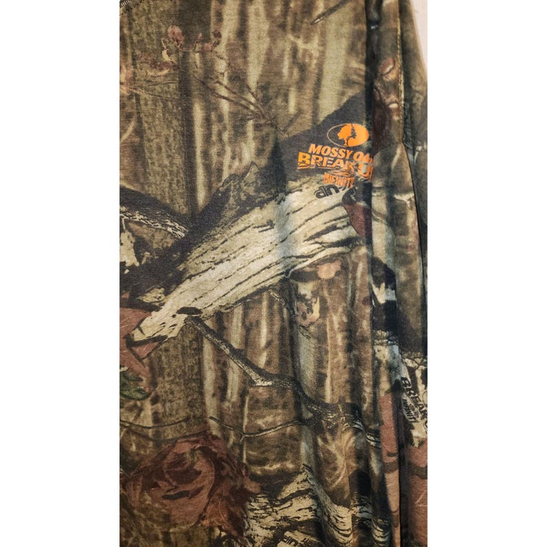 May include: Long-sleeved camouflage shirt with a pattern of brown, green, and tan. The shirt features the "Mossy Oak Break-Up Infinity" logo in orange and white. The fabric appears to be lightweight and suitable for outdoor activities.