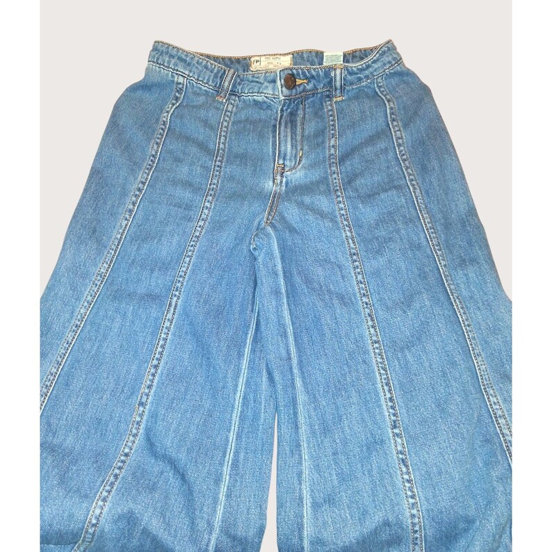 May include: Pale blue denim trousers with a wide leg cut. The trousers have a button and zip fastening, with vertical seams creating a panelled effect. The fabric has a faded wash, giving it a vintage appearance.