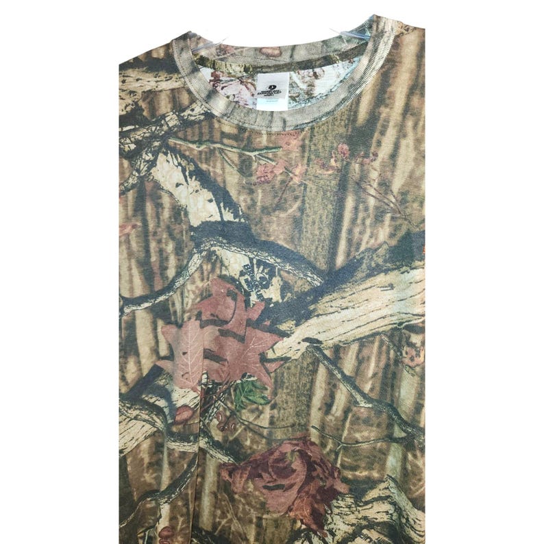 May include: A camouflage-patterned t-shirt in shades of brown, green, and tan. The shirt features a crew neck and a tag visible at the neckline. The pattern includes leaf and branch designs, creating a woodland aesthetic.