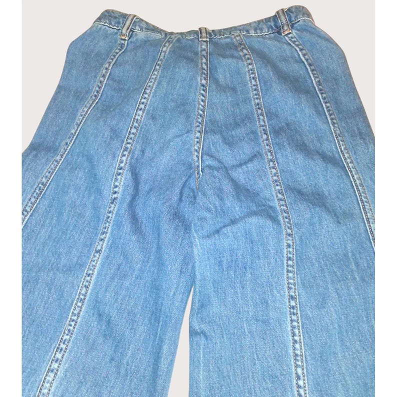 May include: Light blue denim trousers with vertical seam detailing. The trousers have a high waist and a wide leg design. The seams are accented with white stitching. The trousers are made of a medium-weight denim fabric.