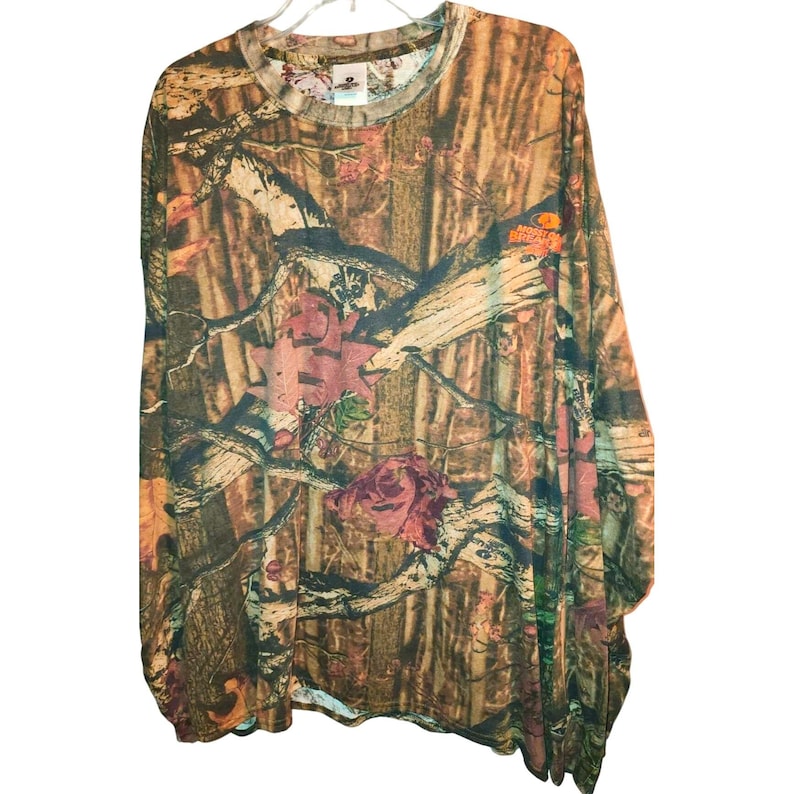 May include: Long-sleeved camouflage shirt in shades of brown, green, and orange. The shirt features a crew neck and a pattern of branches, leaves, and floral elements. The shirt is made of a lightweight material.