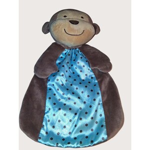 May include: A plush monkey-shaped baby blanket. The monkey has a tan face, brown ears, and a smiling expression. The blanket is brown with a blue satin center featuring black polka dots. A soft and cuddly baby item.