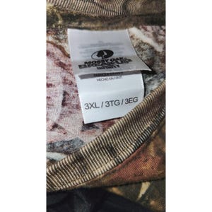 May include: Close-up of a camouflage pattern shirt with a tag. The tag reads "Mossy Oak Break-Up Infinity" and "3XL/3TG/3EG". The shirt is made in Haiti.