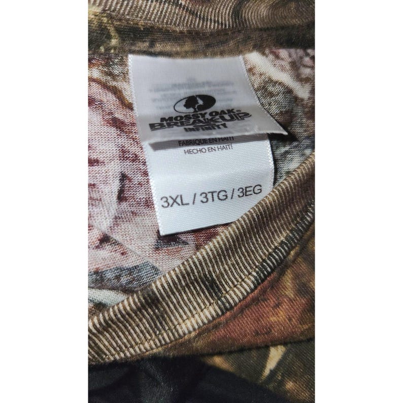May include: Close-up of a camouflage pattern shirt with a tag. The tag reads "Mossy Oak Break-Up Infinity" and "3XL/3TG/3EG". The shirt is made in Haiti.