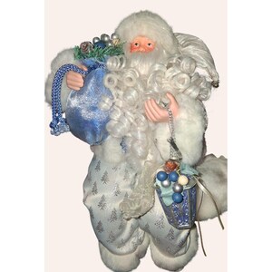 May include: A decorative Santa Claus figurine in a white and silver outfit, holding a blue velvet sack and a blue and silver bucket. The Santa has a white beard and curly hair, with a rosy complexion.