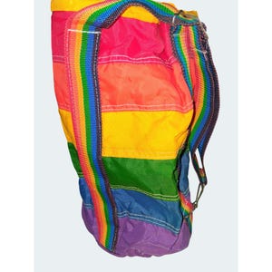 May include: A colorful rainbow-striped backpack with a matching rainbow strap. The bag is cylindrical with a drawstring closure and a rainbow-striped strap. The bag's colors include red, orange, yellow, green, blue, and purple.