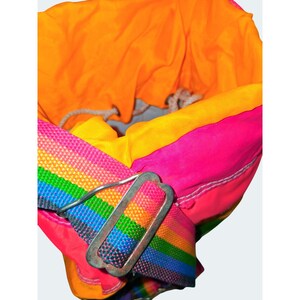 May include: A colorful drawstring bag with a rainbow strap and a silver metal buckle. The bag's interior is orange, with yellow and pink accents. The strap features a vibrant rainbow pattern.