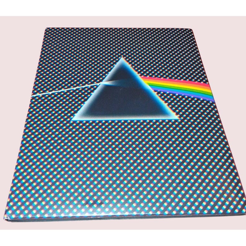 May include: A rectangular notebook featuring a black cover with a repeating pattern of small white dots. Centered on the cover is a large dark triangle with a white light beam entering from the left and exiting as a rainbow spectrum to the right.