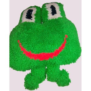 Puede incluir: Un cojín verde con forma de rana con una boca roja sonriente, ojos blancos y pupilas negras. La rana tiene una textura peluda y un diseño caprichoso, ideal para el dormitorio o la sala de juegos de un niño.
