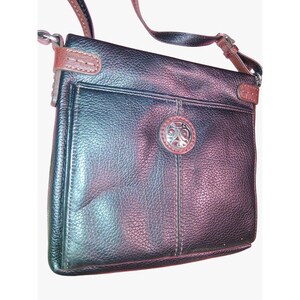 Puede incluir: Un bolso bandolera de cuero negro granulado con ribetes de cuero marr&oacute;n, una correa larga ajustable para el hombro y un emblema met&aacute;lico circular de tono plateado centrado en el bolsillo exterior delantero con detalles en relieve.