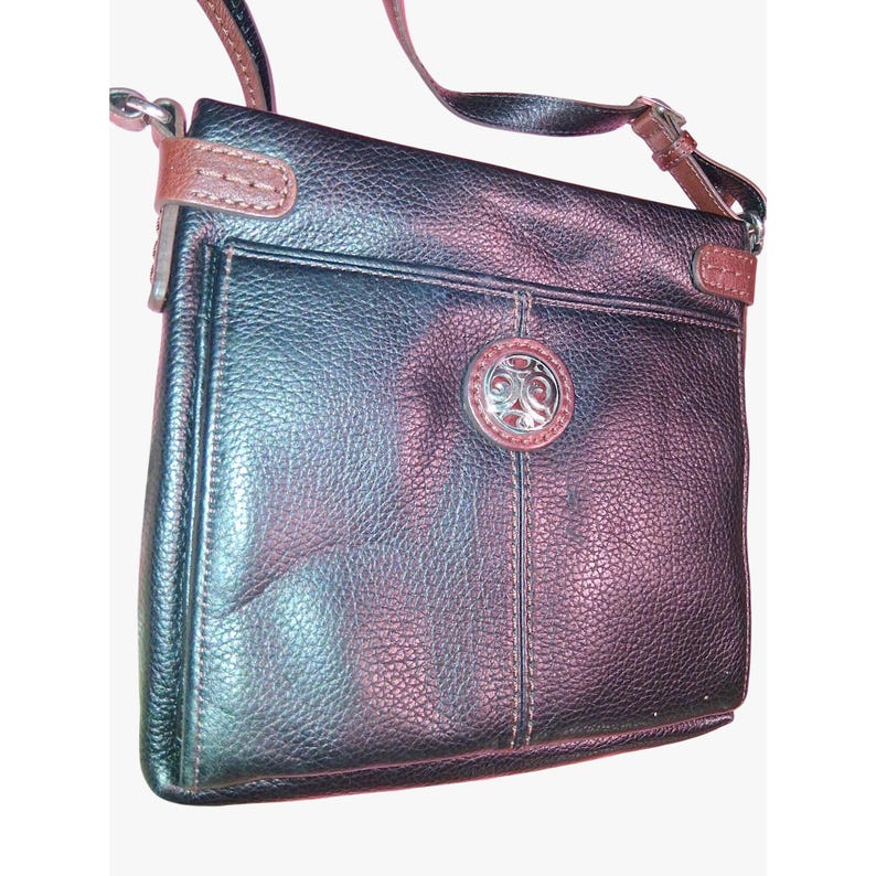 Puede incluir: Un bolso bandolera de cuero negro granulado con ribetes de cuero marr&oacute;n, una correa larga ajustable para el hombro y un emblema met&aacute;lico circular de tono plateado centrado en el bolsillo exterior delantero con detalles en relieve.